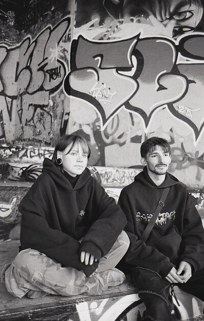 Complices du Bowl Portrait de deux jeunes skateurs, un garçon et une fille, assis et complices devant un mur de graffitis complexes.